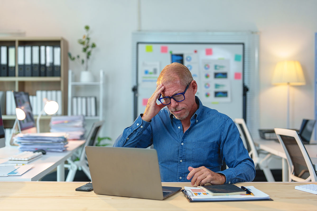 A gray-haired manager in a blue shirt and glasses holds his head in his hand while looking at a laptop at his office desk, with a whiteboard of charts and sticky notes behind him