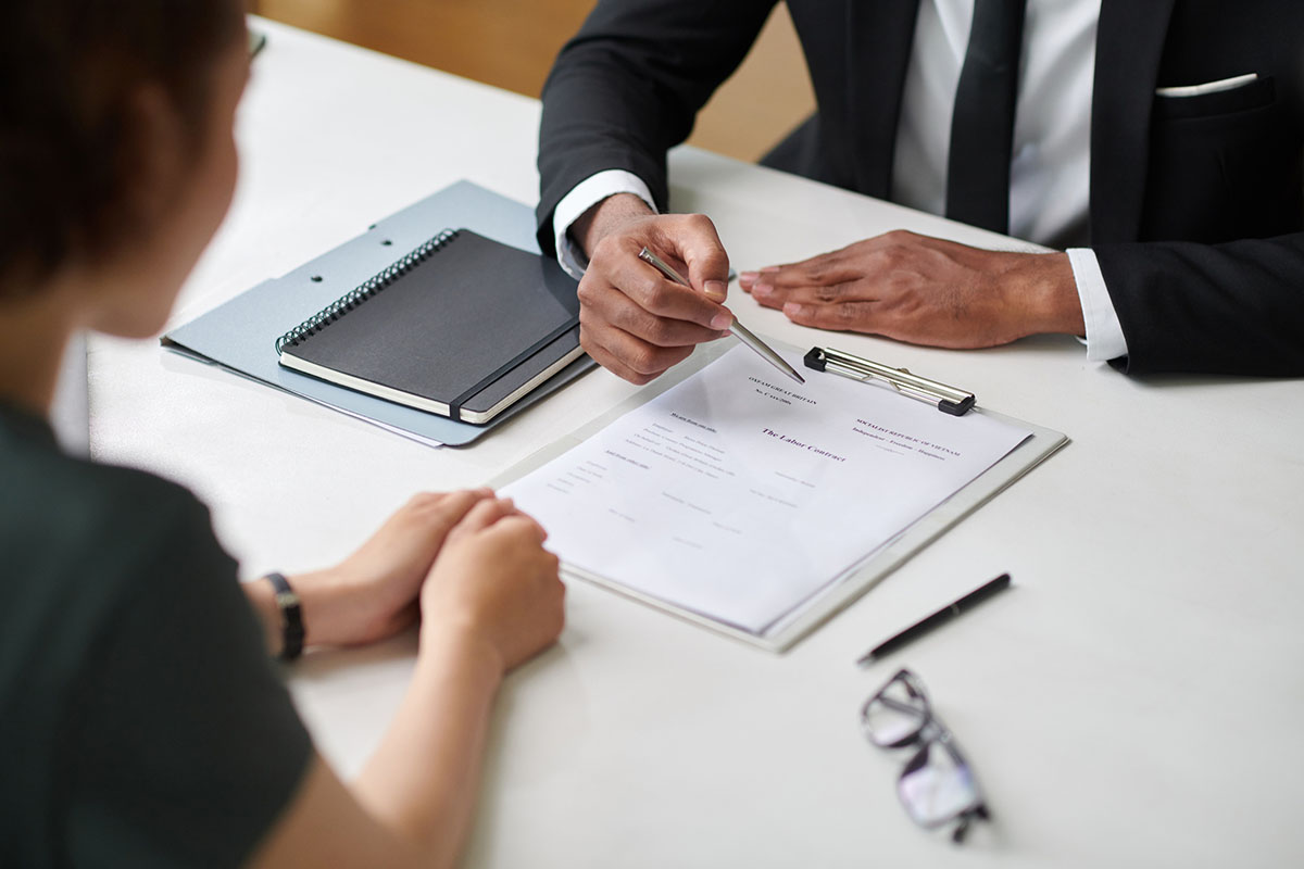 A manager in a dark suit points a pen at an employment contract on a clipboard while a candidate sits across the white desk, with a notebook and glasses nearby