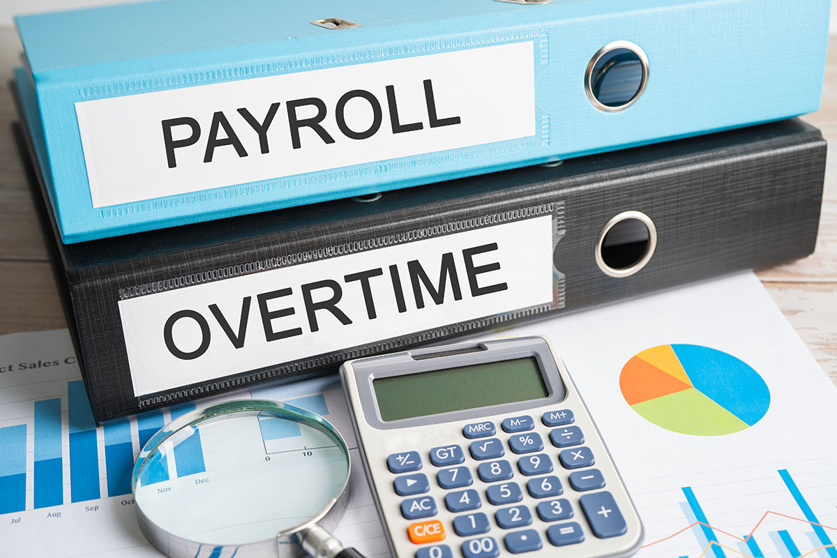 A blue binder labeled Payroll stacked on a black binder labeled Overtime, with a calculator, a magnifying glass and printed bar and pie charts on a desk in front of them
