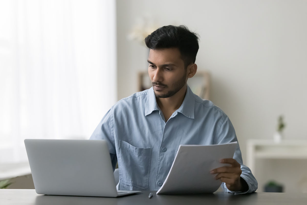 Professional reviewing a printed document alongside a laptop at a desk, with a serious, focused expression