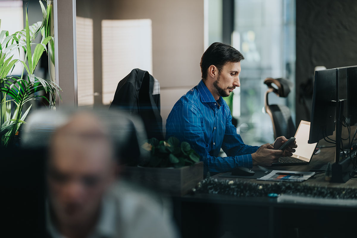 An employee in a blue striped shirt works at a desk with a laptop and phone in a modern office, with a blurred colleague in the foreground