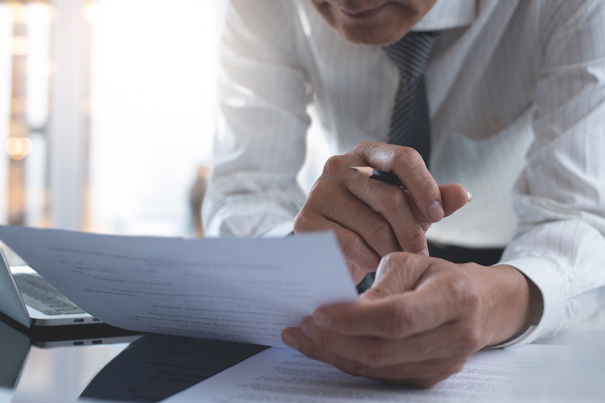 Businessman in a dress shirt and tie closely reviewing the terms of a paper contract at his desk, pen in hand, laptop open beside him
