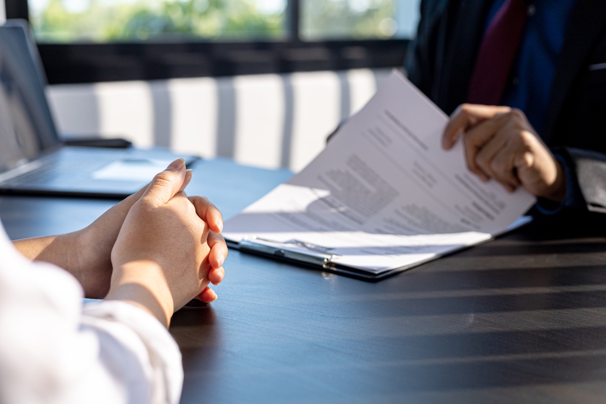 Two people seated across a desk with a printed document between them, one pair of hands folded in the foreground