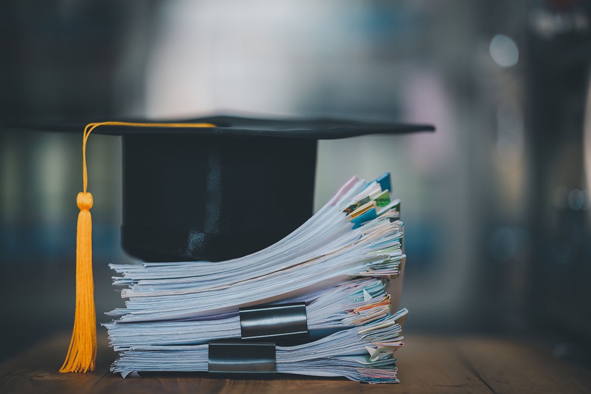 A black graduation cap with a gold tassel resting on a thick stack of clipped paperwork on a wooden desk