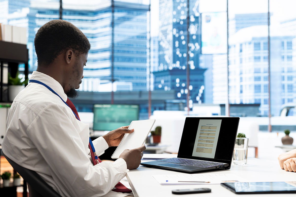 A man in a white shirt and lanyard reads a notebook at an office desk beside an open laptop showing a form, with city towers visible through the window behind him