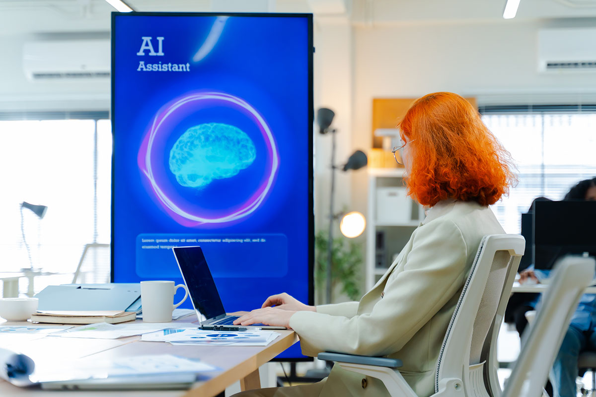 An employee with red hair works on a laptop at an office desk beside a large screen displaying an AI Assistant interface