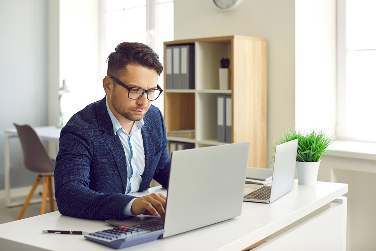 A man in glasses and a navy blazer types on a laptop at a white office desk, with a calculator and pen in front of him and a second laptop and shelf of binders behind