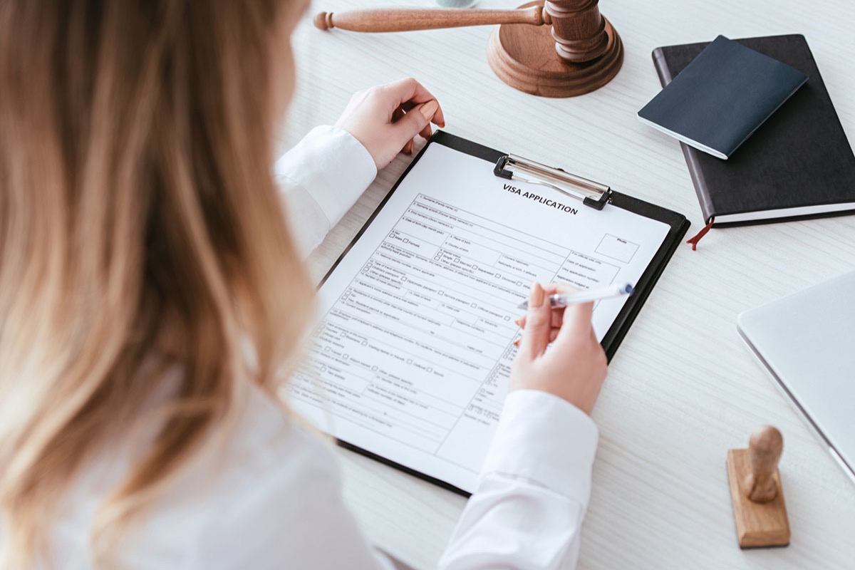 Seen from over her shoulder, a woman fills out a visa application on a clipboard at a white desk beside a judge's gavel, a passport, and a wooden stamp