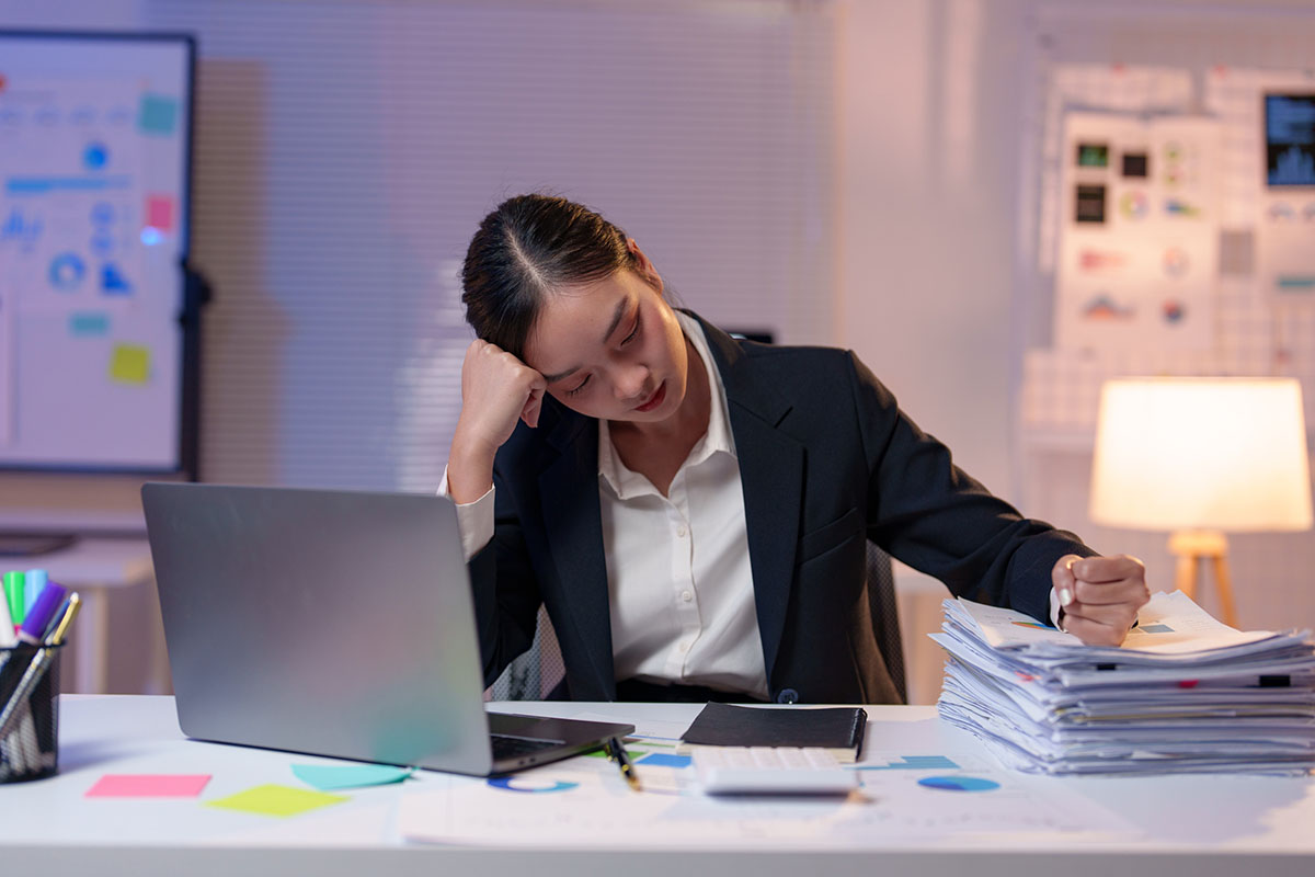 An exhausted professional in a dark blazer rests her head on her hand at an office desk beside a laptop and a tall stack of paperwork