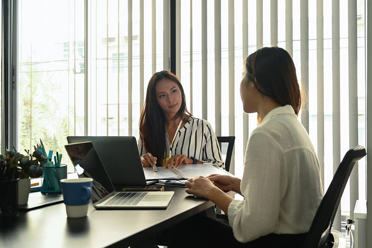 Two colleagues in a one-on-one conversation at an office desk with laptops and an open folder in front of tall windows