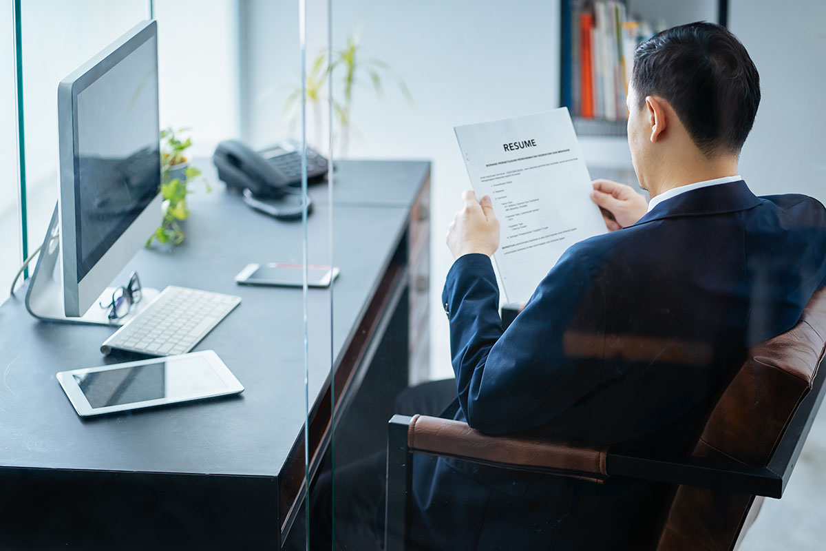 A hiring manager in a navy suit, seen from behind, reading a printed resume at his office desk beside a computer monitor, desk phone, and tablet