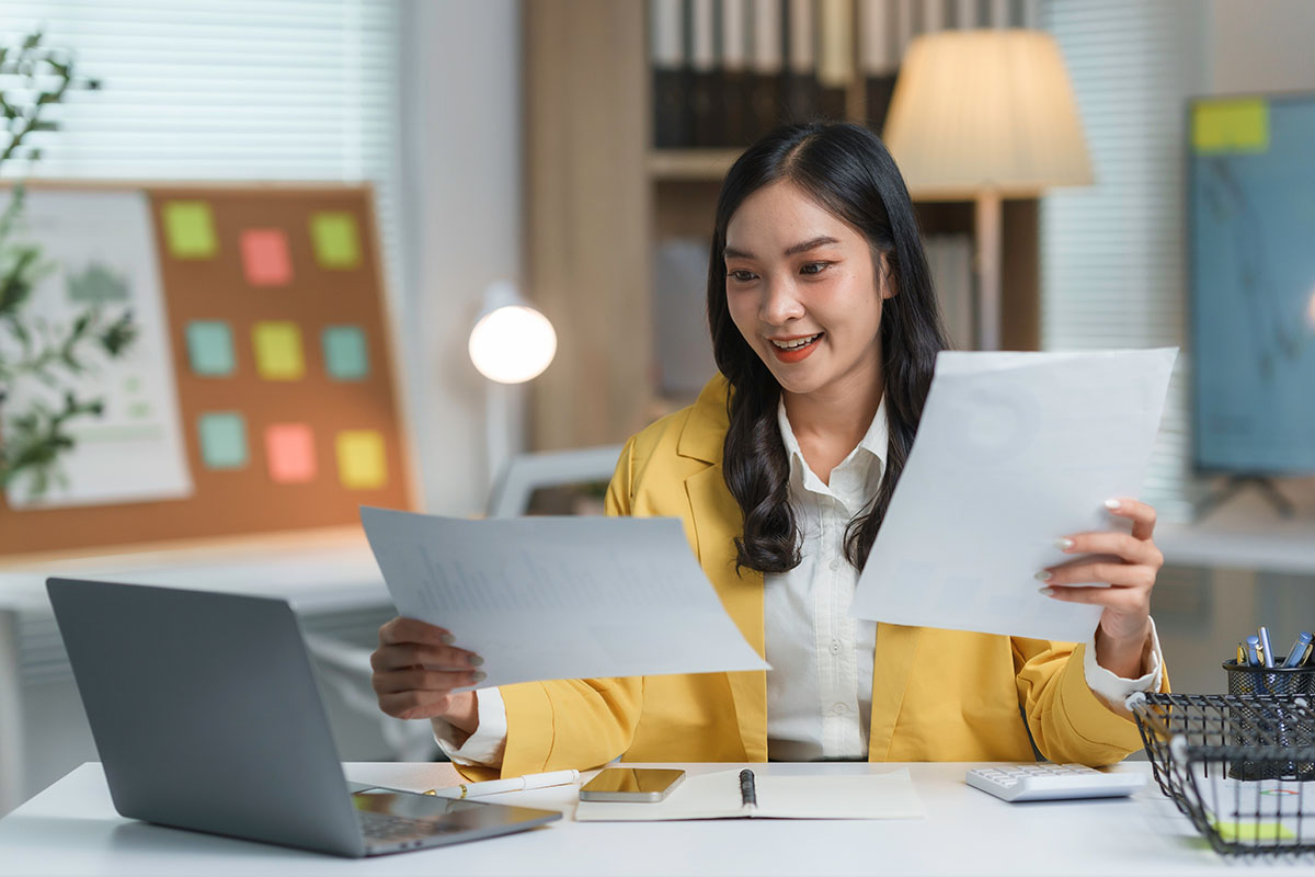 Businesswoman in a modern office comparing two printed reports by hand next to an open laptop