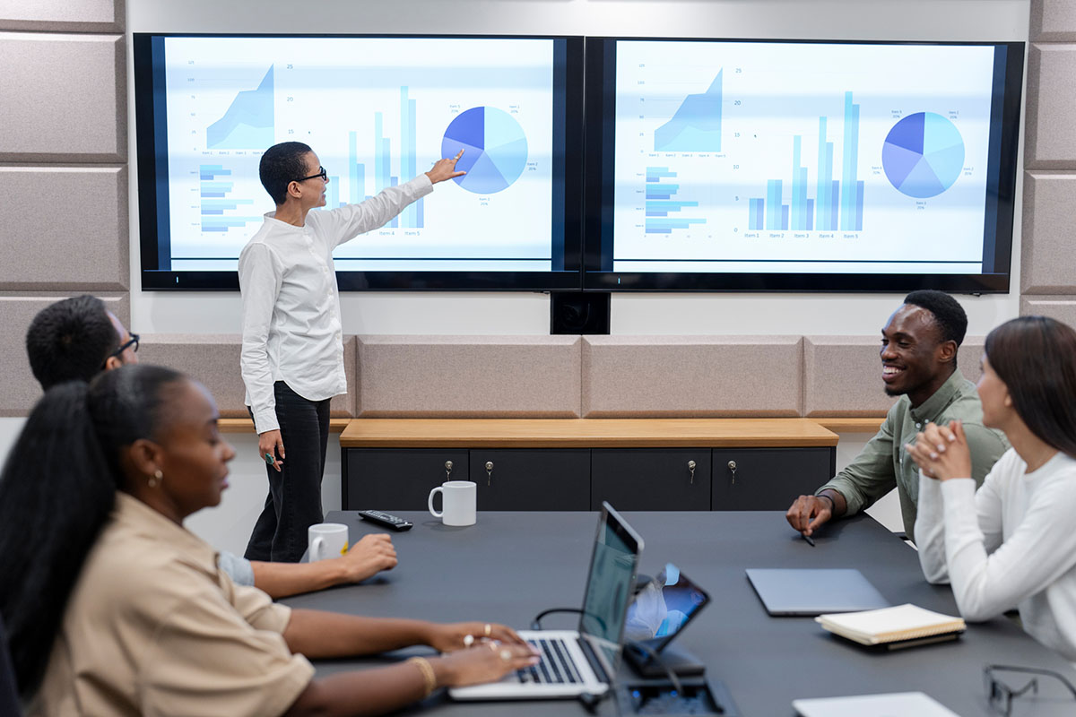 A leader points to bar and pie charts on two large wall screens while colleagues around a conference table with laptops listen