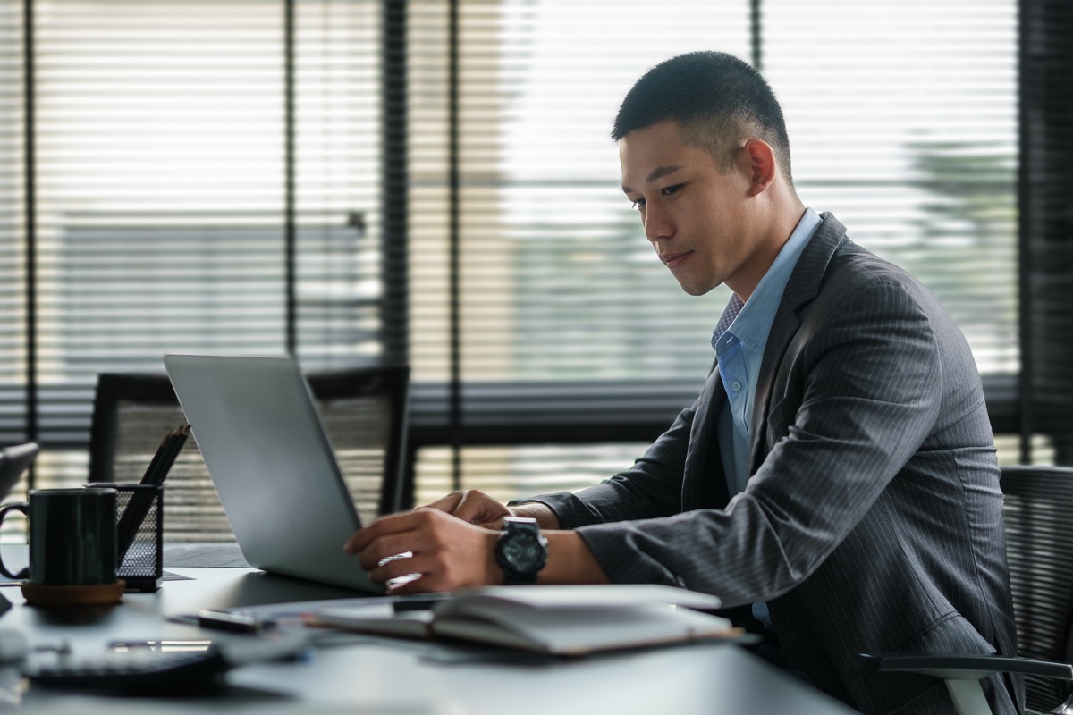 Professional in a gray suit working at a laptop at an office desk, with window blinds behind him