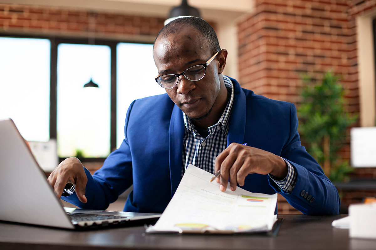 A compensation analyst cross-referencing a pay structure report against a laptop at an office desk