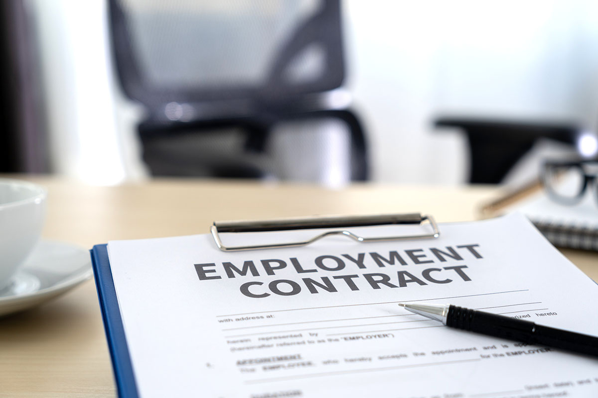 An employment contract on a blue clipboard with a black pen resting across it, on a light wooden desk beside a white coffee cup, with an empty office chair blurred in the background