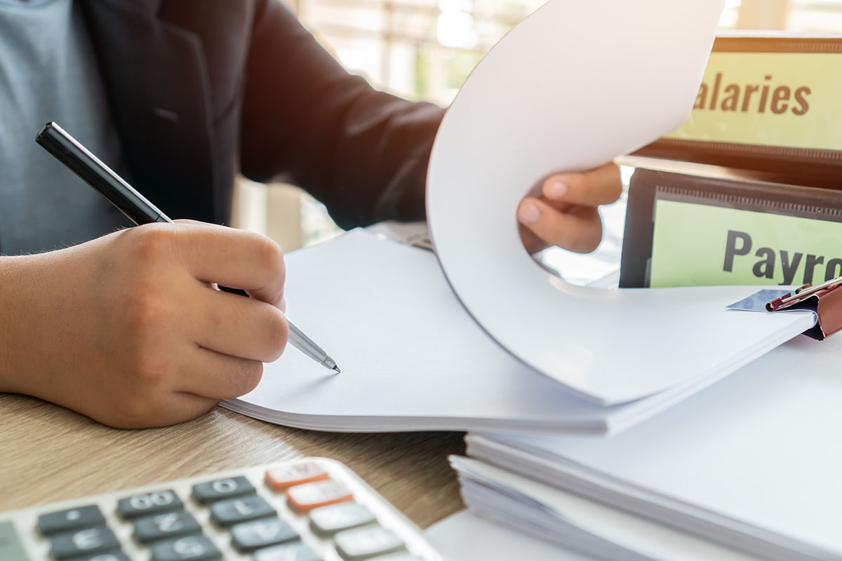 Close-up of a person's hands writing on paperwork next to salary and payroll binders and a calculator on a desk