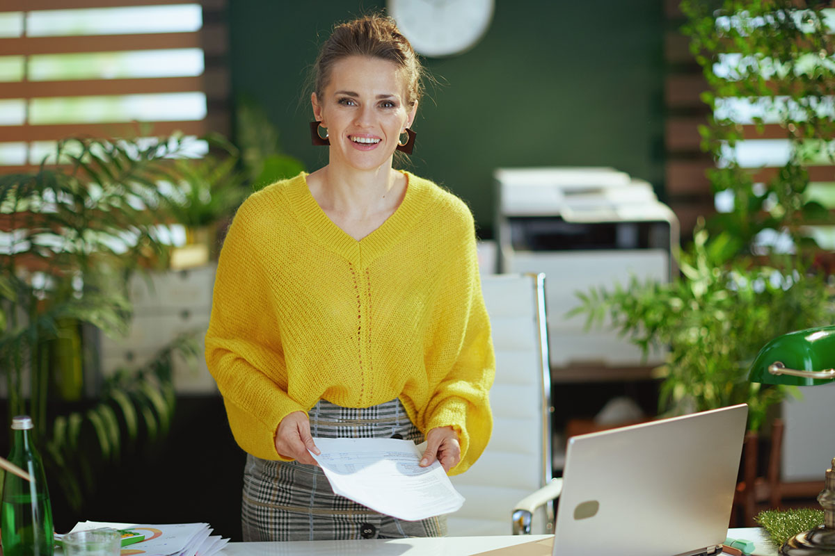 A small business owner in a yellow sweater standing at her office desk holding printed documents beside a laptop