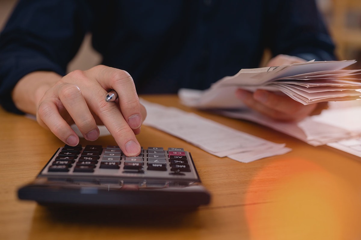 Close-up of a person in a dark shirt tapping a calculator on a wooden desk while holding a stack of receipts and bills