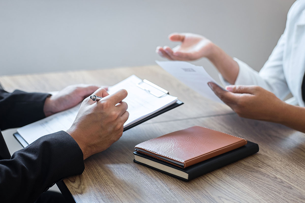 A hiring manager and a candidate reviewing a resume across a wooden desk during an interview, with a clipboard, notebook, and pen visible