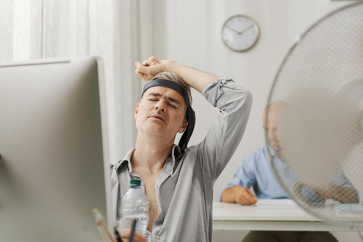 Man at his office desk with a necktie tied around his head, eyes closed in the heat, as a large fan blows and a coworker works behind him
