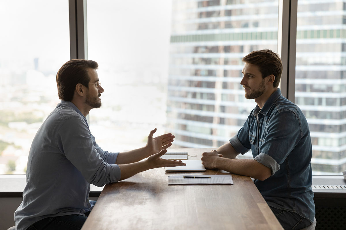 Two men in casual business shirts sit across a long wooden table by a high-rise office window, one gesturing as he explains a point while the other listens, with a document and pen on the table between them