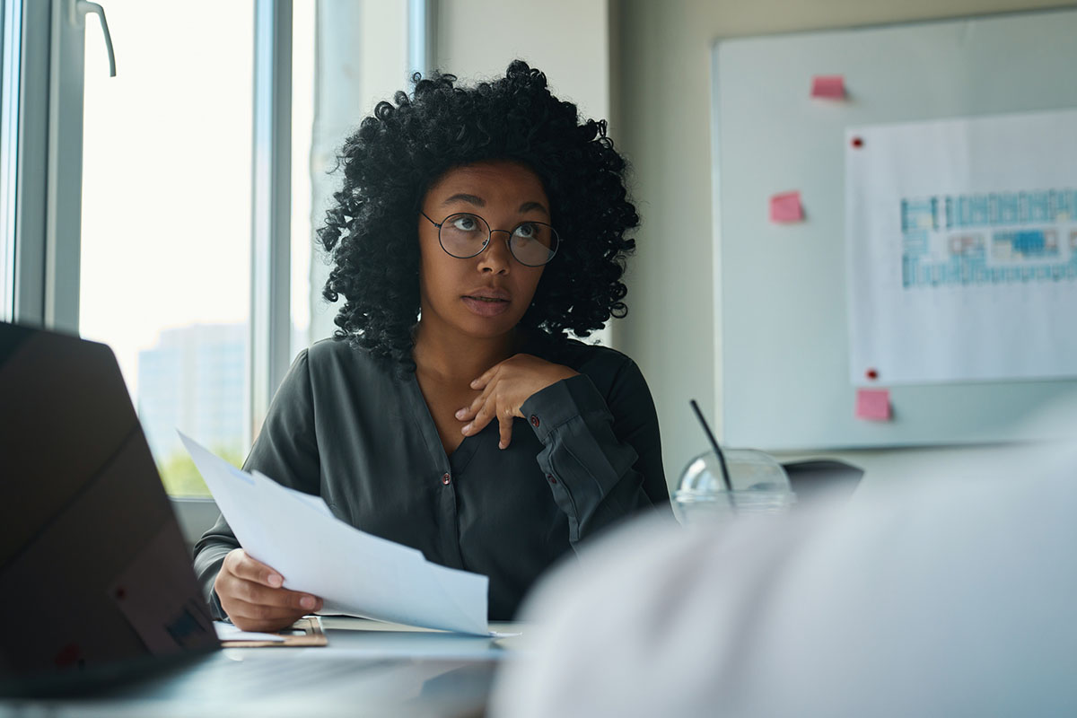 A professional woman in glasses holding printed papers at an office desk, looking up with a skeptical expression