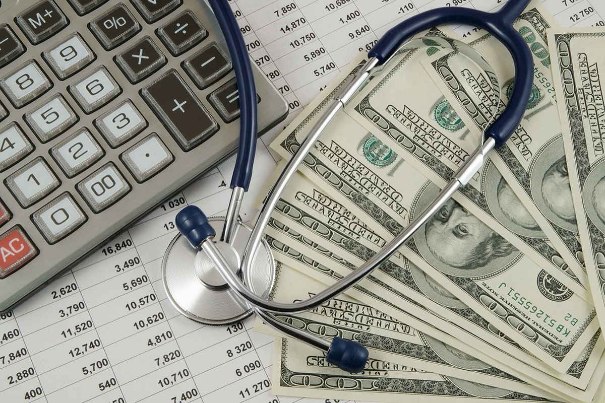 A stethoscope resting on a fan of US dollar bills beside a calculator on a printed cost spreadsheet